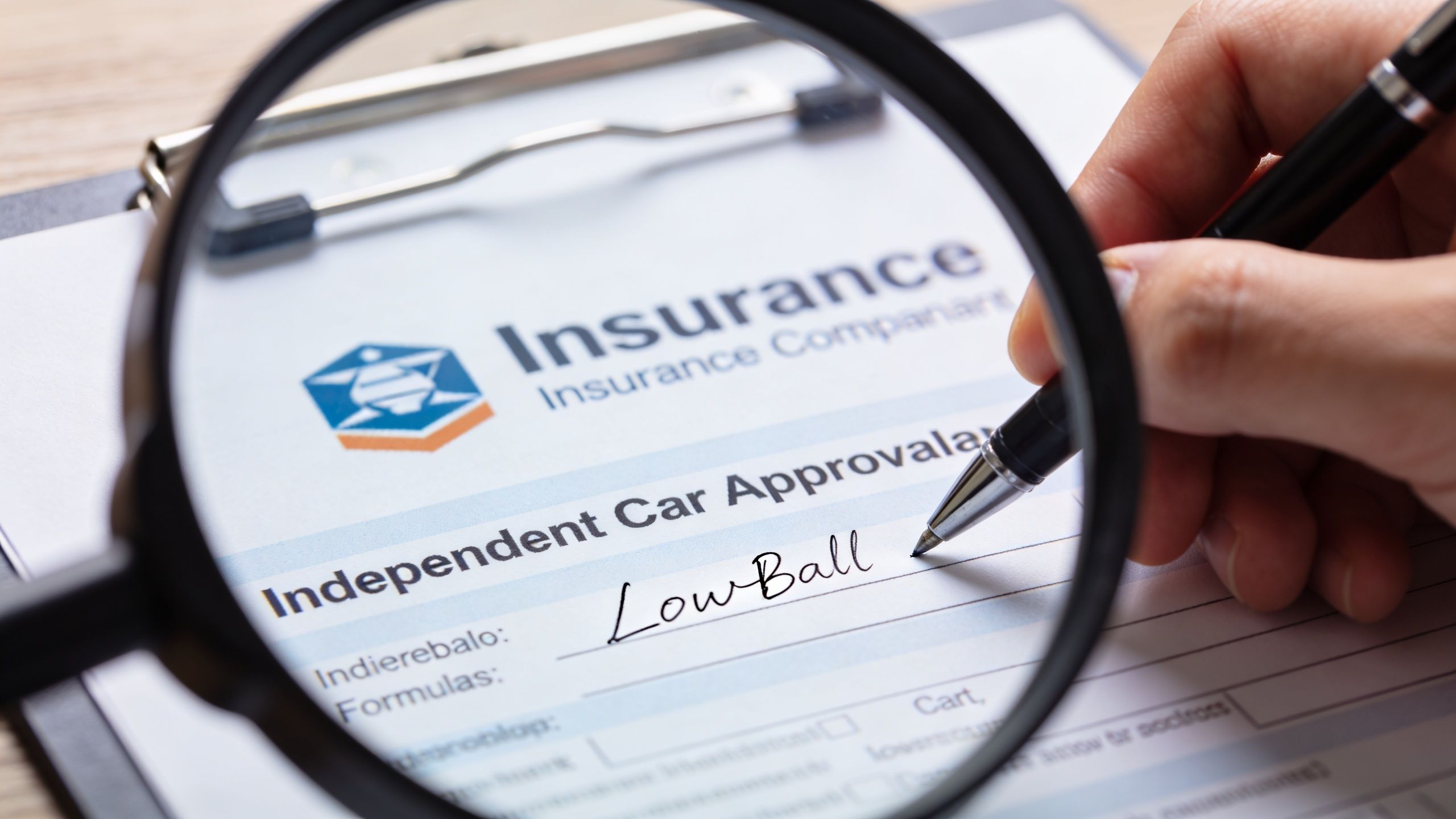 A magnifying glass inspecting an insurance document on a clipboard being signed by a person's hand.
