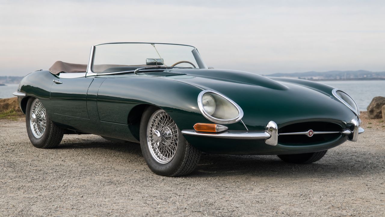 A classic dark green Jaguar E-Type convertible parked on a gravel surface near a calm sea.