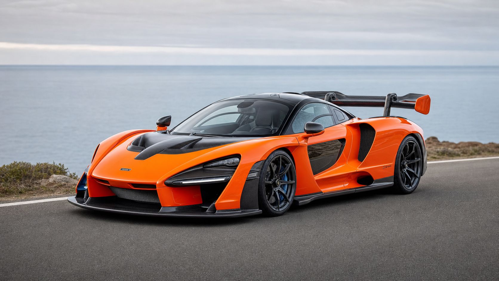 A vibrant orange McLaren Senna hypercar parked on a scenic coastal road overlooking the ocean.